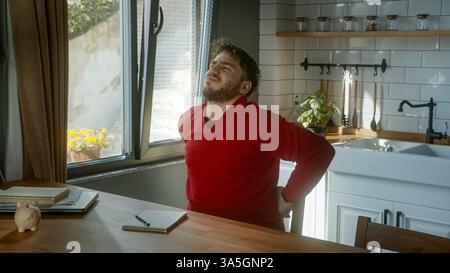 Young man Experiencing Back Pain While Sitting in a Cozy Kitchen Stock Photo