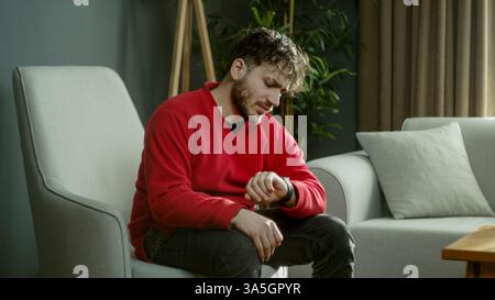 Young man in a red sweater checking the time on his smartwatch while sitting on a couch in a modern living room Stock Photo