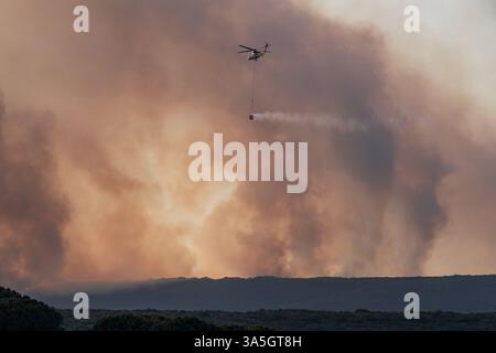 Helicopter drops water over forest fire with large smoke clouds rising in the background. Stock Photo