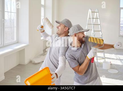 Cheerful male painters singing with paint rollers in bright room, having fun during renovation. Stock Photo