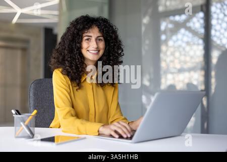 Portrait of a happy successful businesswoman at her workplace inside the office. The woman is smiling and looking at the camera, typing on the laptop keyboard. Stock Photo