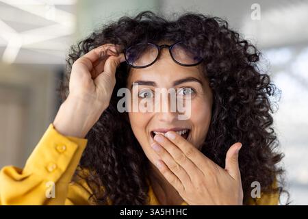 A beautiful woman with curly hair and a yellow blouse, looking surprised as she holds up her glasses indoors, in a close-up portrait Stock Photo