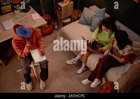 Three college students engaging in research discussion in dorm room setting, with laptop and notepad. Cozy environment with guitar, art supplies and books scattered around Stock Photo