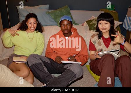 Friends sitting on floor holding notebooks and pens, smiling and posing for camera while studying at home Stock Photo