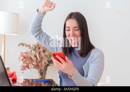 Excited Caucasian woman looking at smartphone while sitting at home office desk Stock Photo