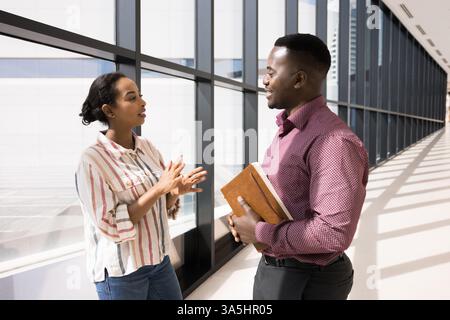 Two African colleagues standing in office hall lead talk, discussing work-related, joint business matters met in contemporary workspace hallway. Busin Stock Photo