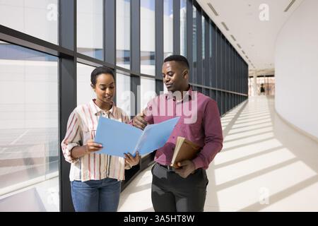 Two young African colleagues standing in modern hallway leading collaborative and professional conversation holds folder reviewing data, discussing de Stock Photo