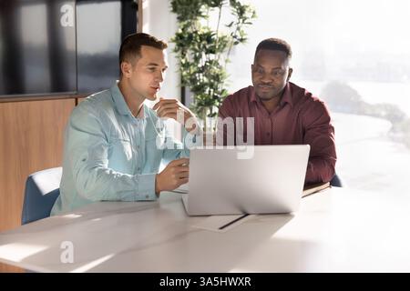 Two young professional men working on joint task using laptop, developing marketing strategy, exchanging point, opinion and vision, gathered in boardr Stock Photo