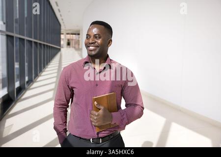 Happy African businessman, male office employee standing in empty hallway looking into distance with dreamy, contented smile, exudes success, competen Stock Photo