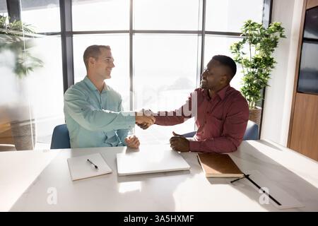 Two young multinational business partners shake hands, start formal meeting, negotiating seated at table in modern in boardroom, smiling, expressing m Stock Photo
