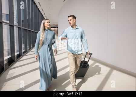 Young couple rolling suitcase moving toward gate. Two Caucasian male, female colleagues walking along modern, well-lit corridor, lead talk, travelling Stock Photo