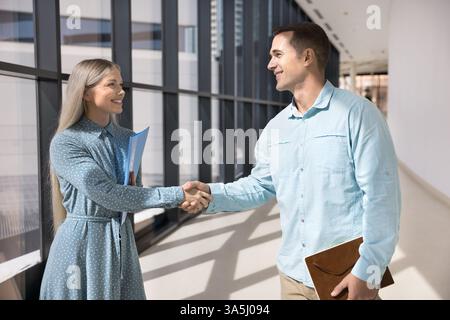 Two smiling European male, female colleagues talking, greeting each other with handshake gesture, met in modern office hallway, express mutual respect Stock Photo