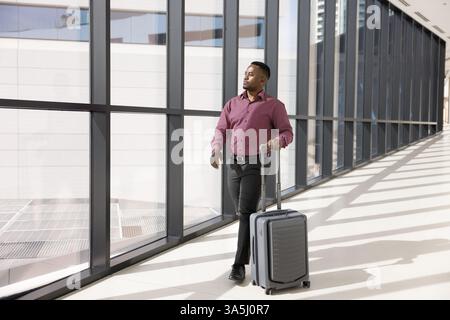 African man dressed in formal shirt and black trousers, walking along hallway with large windows, pulling rolling suitcase, hurrying to catch flight, Stock Photo