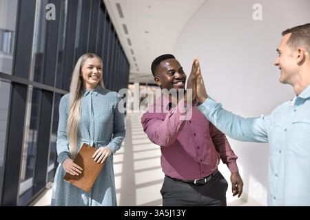 Two young 30s multiethnic male colleagues, buddies welcoming each other, giving high-five gesture during informal meeting in corporate office hallway, Stock Photo