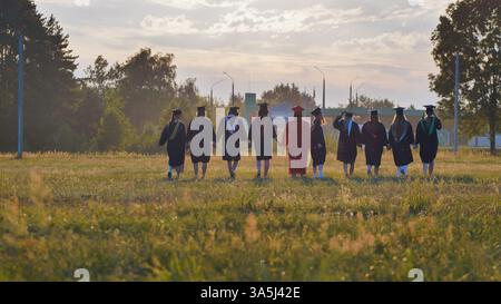 Graduates in cap and gown walking through a field at sunset, celebrating their academic success and camaraderie by holding hands Stock Photo
