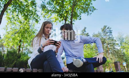 Two high school students are watching a smartphone during the school break sitting on a low wall in a park Stock Photo