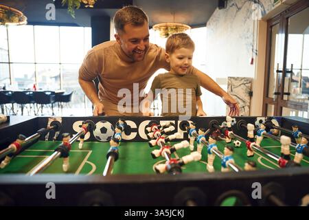 Teaching how to play. Father and son are playing table football game indoors. Stock Photo