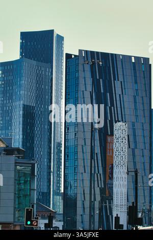 The image shows multiple modern, blue-toned skyscrapers and an ornamental tower in an urban setting, under a pale sky in Manchester, UK. Stock Photo