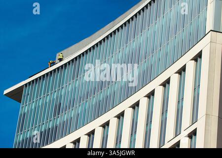 Low-angle shot of a modern building with glass and white concrete, and two workers on the roof, against a clear blue sky in Manchester, UK. Stock Photo
