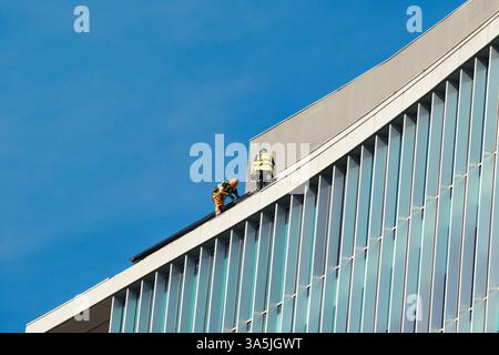 Two workers in safety gear on the roof of a modern building with a glass facade, set against a clear blue sky in Manchester, UK. Stock Photo