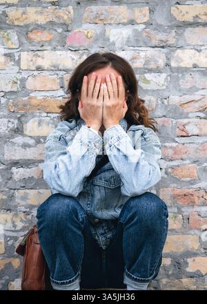 Woman sitting outdoors next to a brick wall, covering her face with her hands in a gesture of distress or sadness. Stock Photo