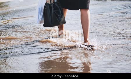 Businesswoman wading through dirty flood water on city street after heavy rain, holding bag and wearing black shoes Stock Photo