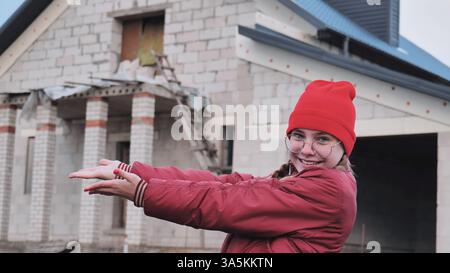 Female architect presenting house blueprint, pointing confidently while standing near construction site model Stock Photo