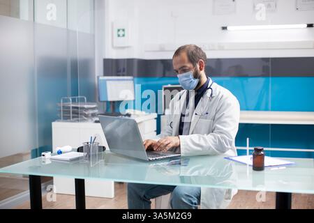 Male general practitioner using laptop for reviewing medical records and checking emails in hospital room. Masked doctor sitting at table with protective screen preparing for patient appointments. Stock Photo