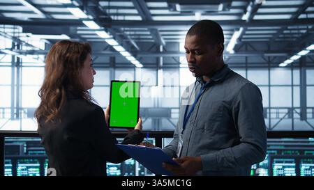 Computer scientists in data center use mockup tablet, inspecting server rigs, updating devices. Teamworking IT colleagues collaborating to ensure stable operations, using chroma key device, camera B Stock Photo