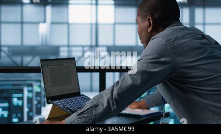 Technician coding on laptop in data center, managing server virtualization, finetuning network protocols. Server farm employee programming on notebook, enforcing advanced security measures, camera A Stock Photo