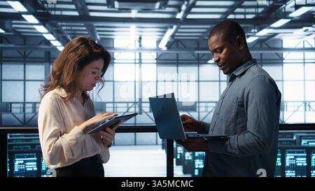 Developers in data center using laptop and tablet, executing security patches, securing firewall. Server farm workers using devices to perform vulnerability scans, ensuring data protection, camera B Stock Photo