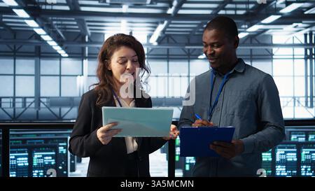 Data center management executives reading paperwork files, reviewing system diagnostics. Server farm colleagues looking over maintenance schedules documents and cybersecurity protocols, camera B Stock Photo
