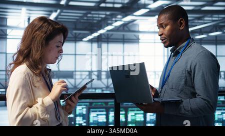 Team of technicians working in server farm, using laptops to analyze data. Teamworking workers examining infrastructure in data center, ensuring system integrity and security, camera B Stock Photo