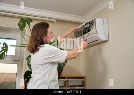 Woman repairing wall-mounted air conditioner with screwdriver in a home interior. HVAC maintenance and repair concept. Stock Photo