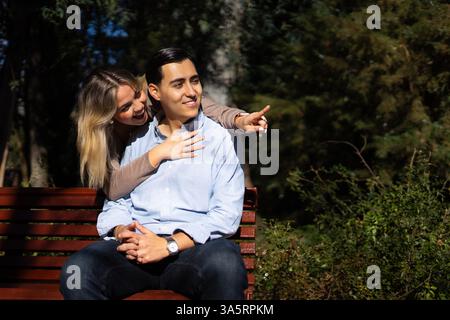 Young latin couple sitting on a bench in a park, pointing at something interesting Stock Photo