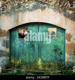 The worn green doors, adorned with aged wood and broken glass, reflect time's passage in a serene rural setting. Auvergne. France Stock Photo