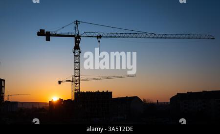 Cranes tower over a construction site, silhouetted by the glowing orange and purple hues of the sunset. The skyline features buildings gradually being Stock Photo