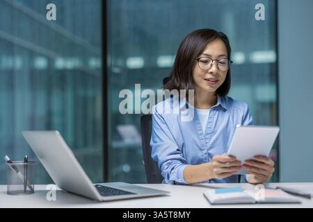 Young adult Asian woman in glasses works on her tablet at her desk in an office. Laptop and notepad are also on the desk. Stock Photo