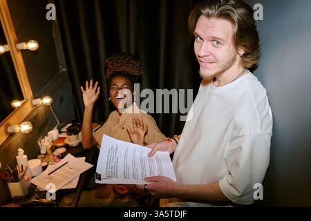 Young man holding script and smiling at camera while young woman waves sitting in dressing room Collaborating and preparing for an upcoming event or performance Stock Photo