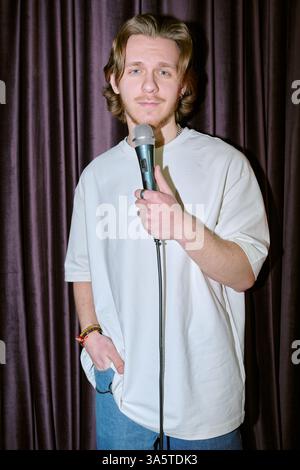 Portrait of young man with long hair standing in front of dark curtain, holding microphone, wearing casual clothes, smiling directly at camera, appearing confident Stock Photo