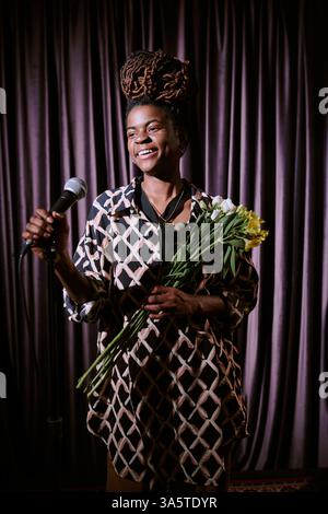 Woman with braided hair smiling while holding microphone and bouquet of flowers on stage with curtain backdrop. Performing or presenting with bright expression Stock Photo