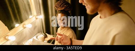 African American woman with braids smiling while listening attentively in dressing room with bright lights and mirrors Stock Photo