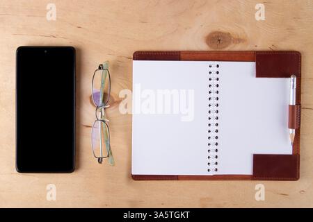 Digitalization concept. Paper notebook vs smartphone.  Eyeglasses, Digital phone with blank black screen and empty paper pad lie on wooden desk Stock Photo