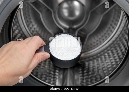Cleaning washing machine drum with Citric acid. Person hand holding measuring cup filled with citric acid against washing machine drum in home indoors Stock Photo