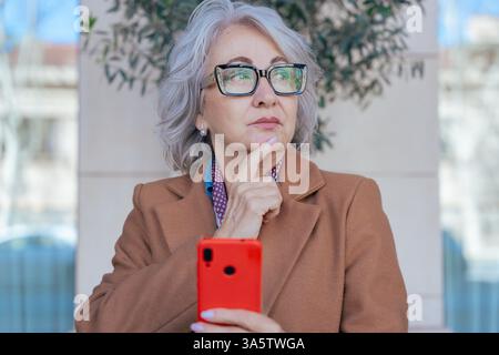 Thoughtful senior woman with smartphone looking away in an urban environment Stock Photo
