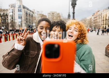 Three multi-ethnic female tourists are laughing and taking a selfie with a smartphone, one of them is making the victory sign, in a square in barcelon Stock Photo