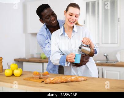 Happy biracial couple laughing and drinking coffee in morning in kitchen Stock Photo
