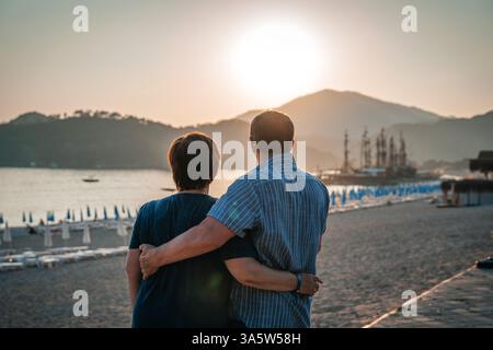 Two elderly men stand on the beach with their arms around each other watching the sunset over the sea. Mountains and ships can be seen in the distance Stock Photo