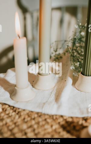 A table with three candles, one of which is lit. The other two are unlit. The candles are placed on a white cloth, and there are some flowers in the b Stock Photo