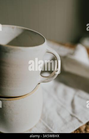 Two white ceramic mugs are stacked on top of each other on a white cloth. The mugs are plain and simple, with no designs or patterns on them. Concept Stock Photo